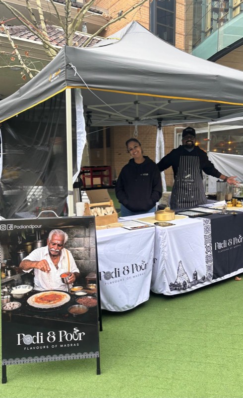 The Podi & Pour stall on the green mat — a white marquee, branded tables and the A-stand poster out front
